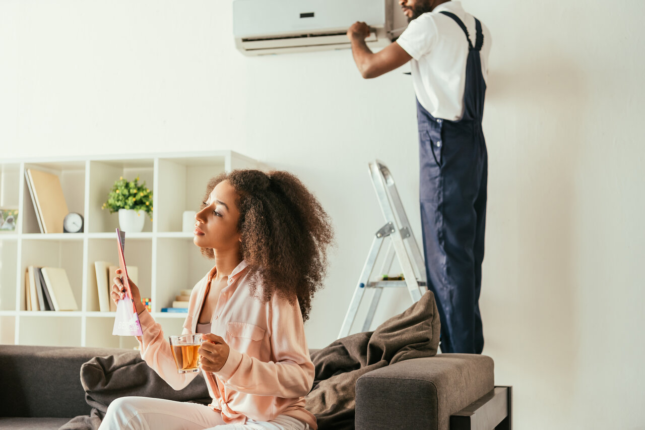 HVAC Blog 5 Thoughtful African American woman sitting on sofa holding a cup of tea, while HVAC technician services air conditioning unit on wall in cozy living room setting.