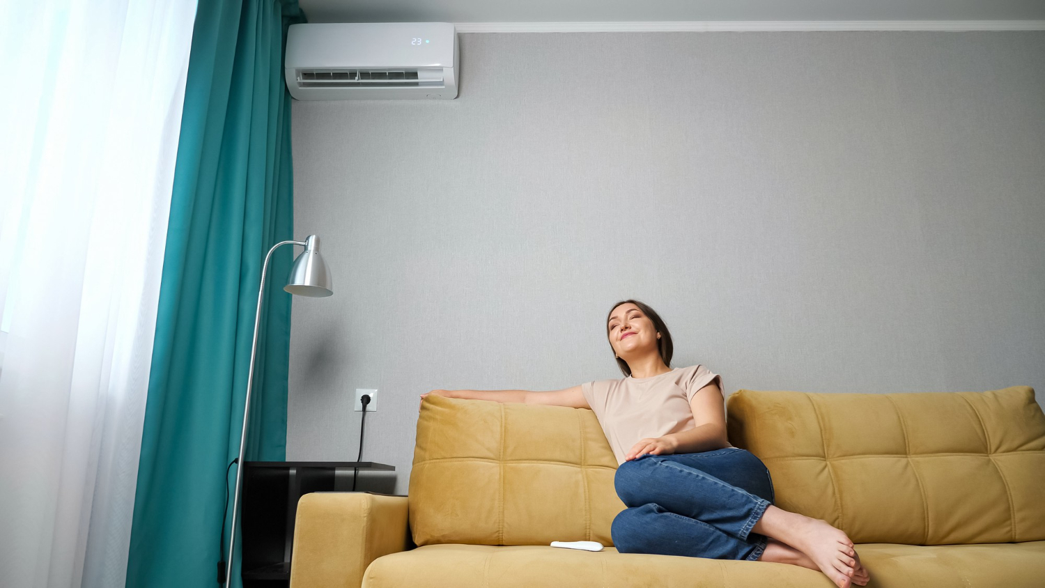 HVAC Blog 3 Woman relaxing on a yellow couch in a living room with an air conditioning unit, enjoying cool air during summer.