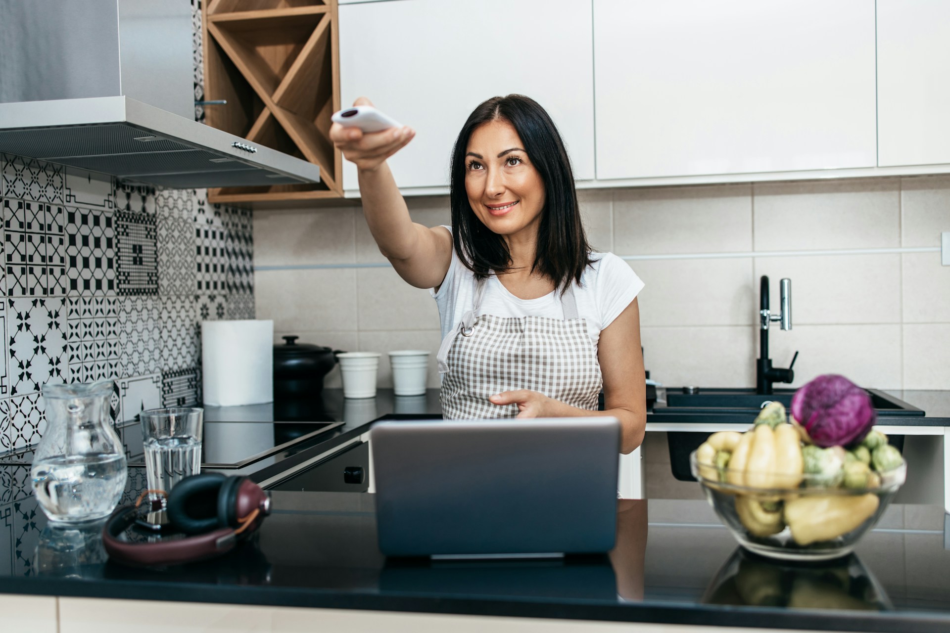 HVAC Blog 6 Woman in a kitchen using a remote control while standing near a laptop and a bowl of fresh vegetables, illustrating home comfort and technology in HVAC discussions.