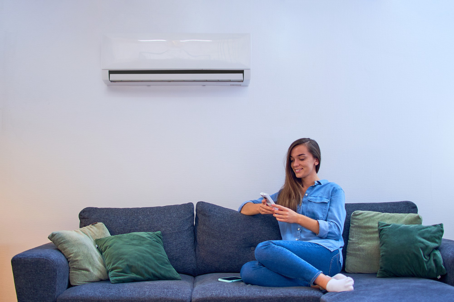 HVAC Blog 9 Woman sitting on a couch using a remote control for an air conditioning unit mounted on the wall, surrounded by green pillows, illustrating comfort and HVAC efficiency.