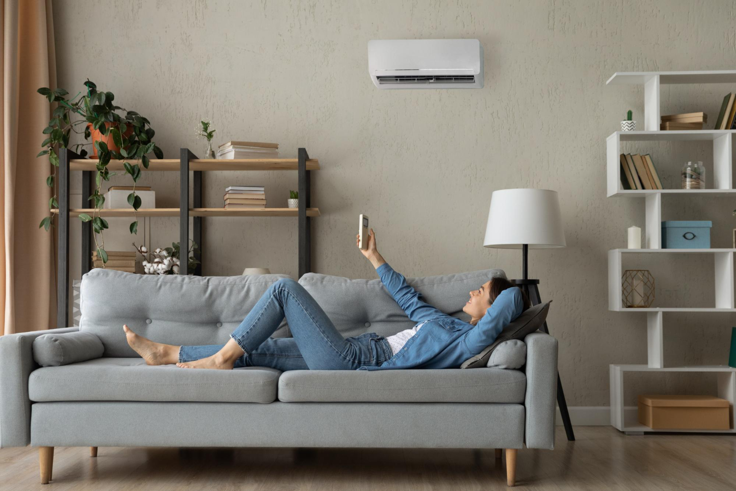 HVAC Blog 8 Woman relaxing on a gray sofa using a smartphone, with an air conditioning unit mounted on the wall, surrounded by indoor plants and bookshelves, illustrating comfortable home living in relation to HVAC services.